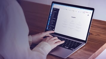 Eine Person sitzt an einem Holztisch und verwendet einen Laptop mit der ChatGPT-Seite im Browser.