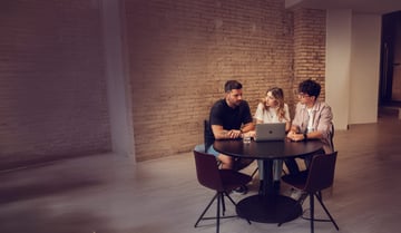 Drei Personen sitzen an einem runden Tisch in einem Raum mit einer unverputzten Ziegelwand und arbeiten gemeinsam an einem Laptop.