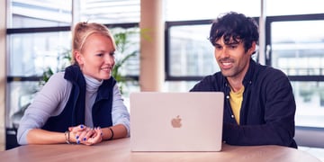 Zwei Personen arbeiten gemeinsam an einem Laptop in einem hellen und modernen Büroraum mit großen Fenstern.